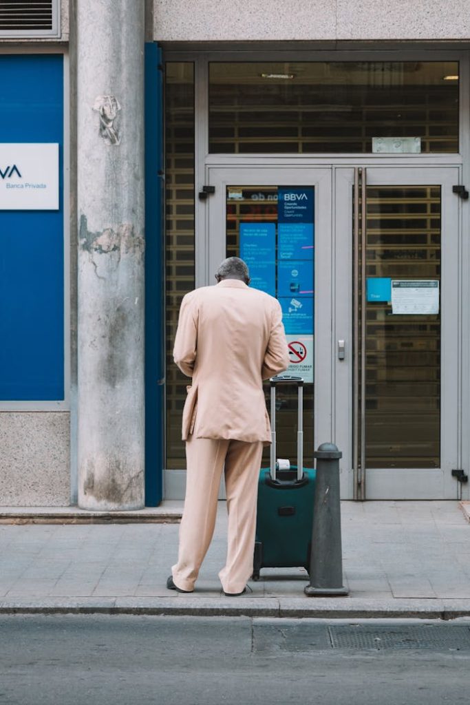pexels-photo-17976144 Man in a suit with luggage standing outside a bank in an urban setting